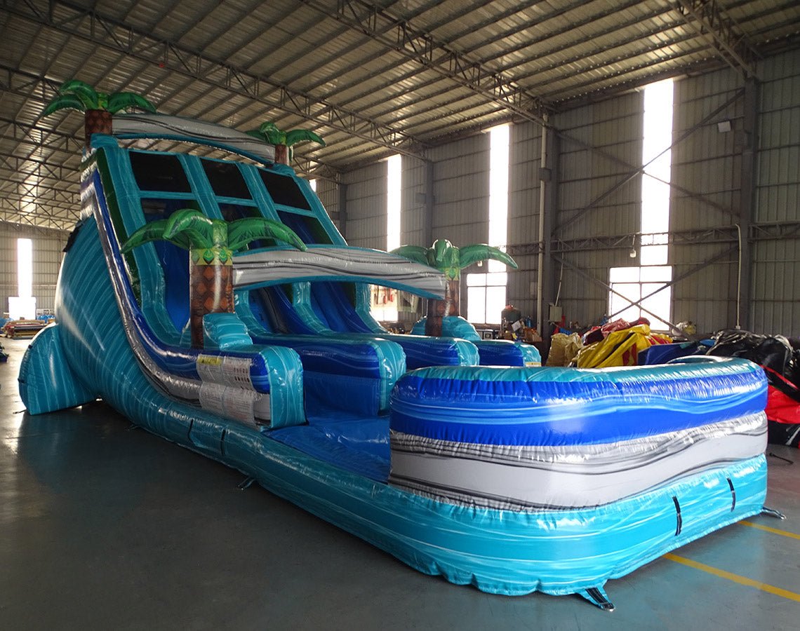 Three-quarter view of 18ft Bahama Blast water slide displaying blue lanes, tropical-themed design with palm trees, central climbing structure, and large splash area surrounded by other inflatable products in a warehouse.