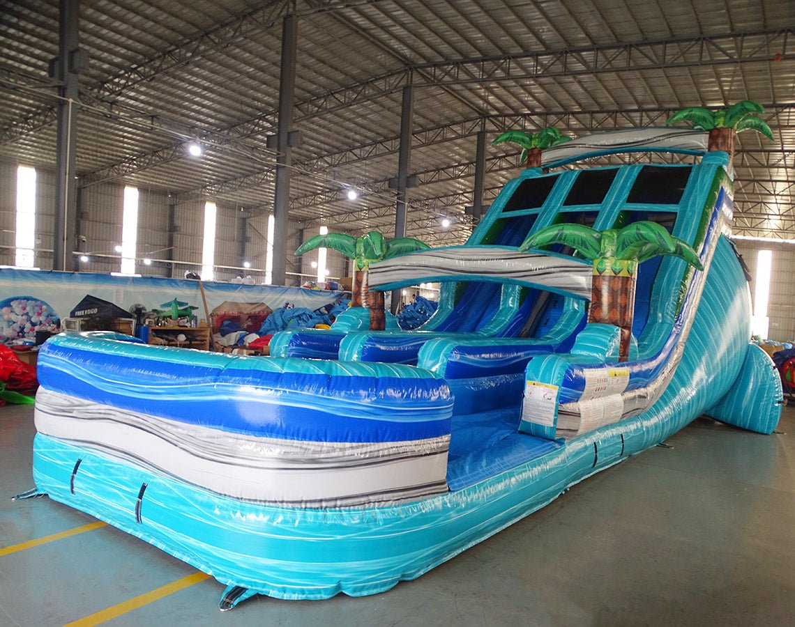 Angled view of 18ft Bahama Blast Center Climb water slide showcasing its full length, three blue sliding lanes, inflatable palm tree decorations, central climbing section, and extended splash pool in an indoor facility.