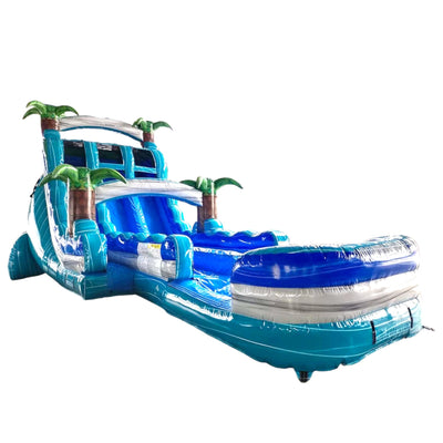 Large inflatable water slide with tropical theme, featuring a tall turquoise and blue structure with palm tree decorations, dual sliding lanes, and an extended splash pool.