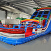 Three-quarter view of 18ft Baja Splash water slide displaying blue lanes, tropical-themed design with palm trees, rainbow arches, central climbing structure, and large red and blue splash area in a warehouse surrounded by other inflatables.