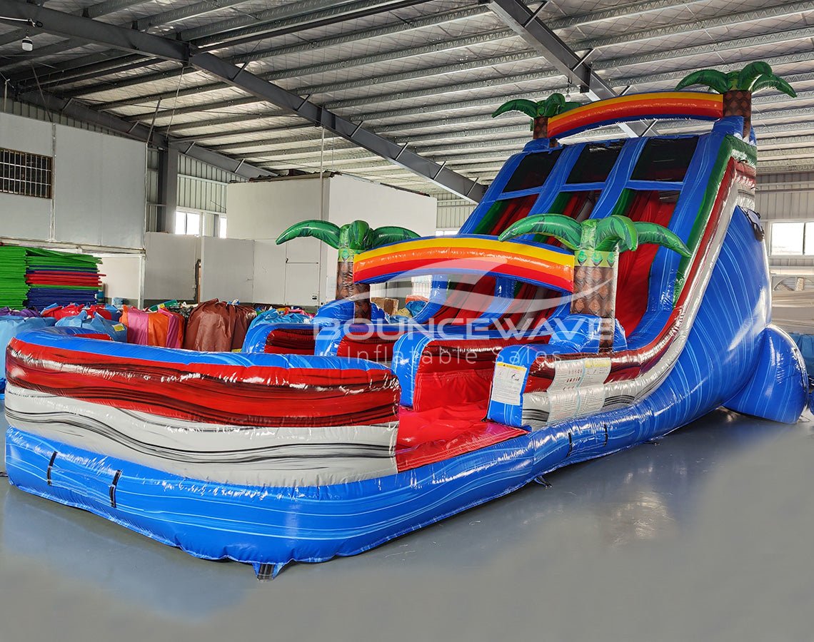 Three-quarter view of 18ft Baja Splash water slide displaying blue lanes, tropical-themed design with palm trees, rainbow arches, central climbing structure, and large red and blue splash area in a warehouse surrounded by other inflatables.