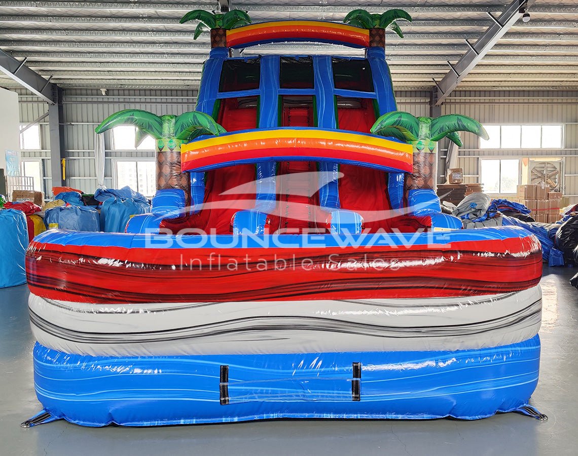 Close-up front view of 18ft Baja Splash Center Climb water slide showcasing the vibrant blue, red, and white color scheme, inflatable palm tree decorations, rainbow arches, and detailed wave patterns on the splash pool.