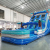 Three-quarter view of 18ft Cayman Crush water slide displaying blue lanes, tropical-themed design with palm trees, central climbing structure, and large turquoise splash area in an indoor facility with other inflatables visible.