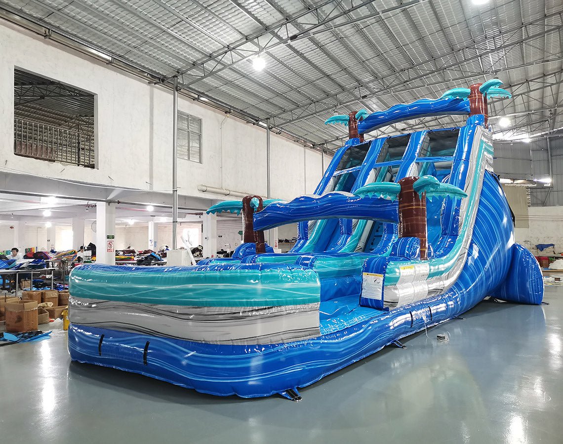 Three-quarter view of 18ft Cayman Crush water slide displaying blue lanes, tropical-themed design with palm trees, central climbing structure, and large turquoise splash area in an indoor facility with other inflatables visible.