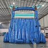 Large inflatable water slide featuring a tropical theme with blue wave-patterned lanes, turquoise palm tree decorations, brown inflatable tree trunks, and a spacious turquoise splash pool, set up in an indoor warehouse with visible manufacturing equipment.