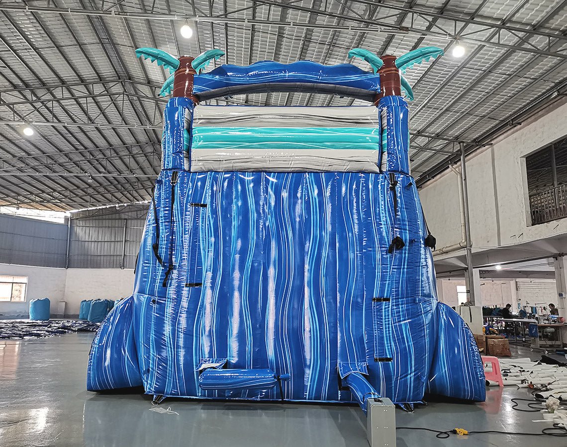 Large inflatable water slide featuring a tropical theme with blue wave-patterned lanes, turquoise palm tree decorations, brown inflatable tree trunks, and a spacious turquoise splash pool, set up in an indoor warehouse with visible manufacturing equipment.