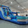 Angled view of 18ft Cayman Crush Center Climb water slide showcasing its full length, three blue sliding lanes, inflatable palm tree decorations, central climbing section, and extended turquoise splash pool in a large indoor warehouse.