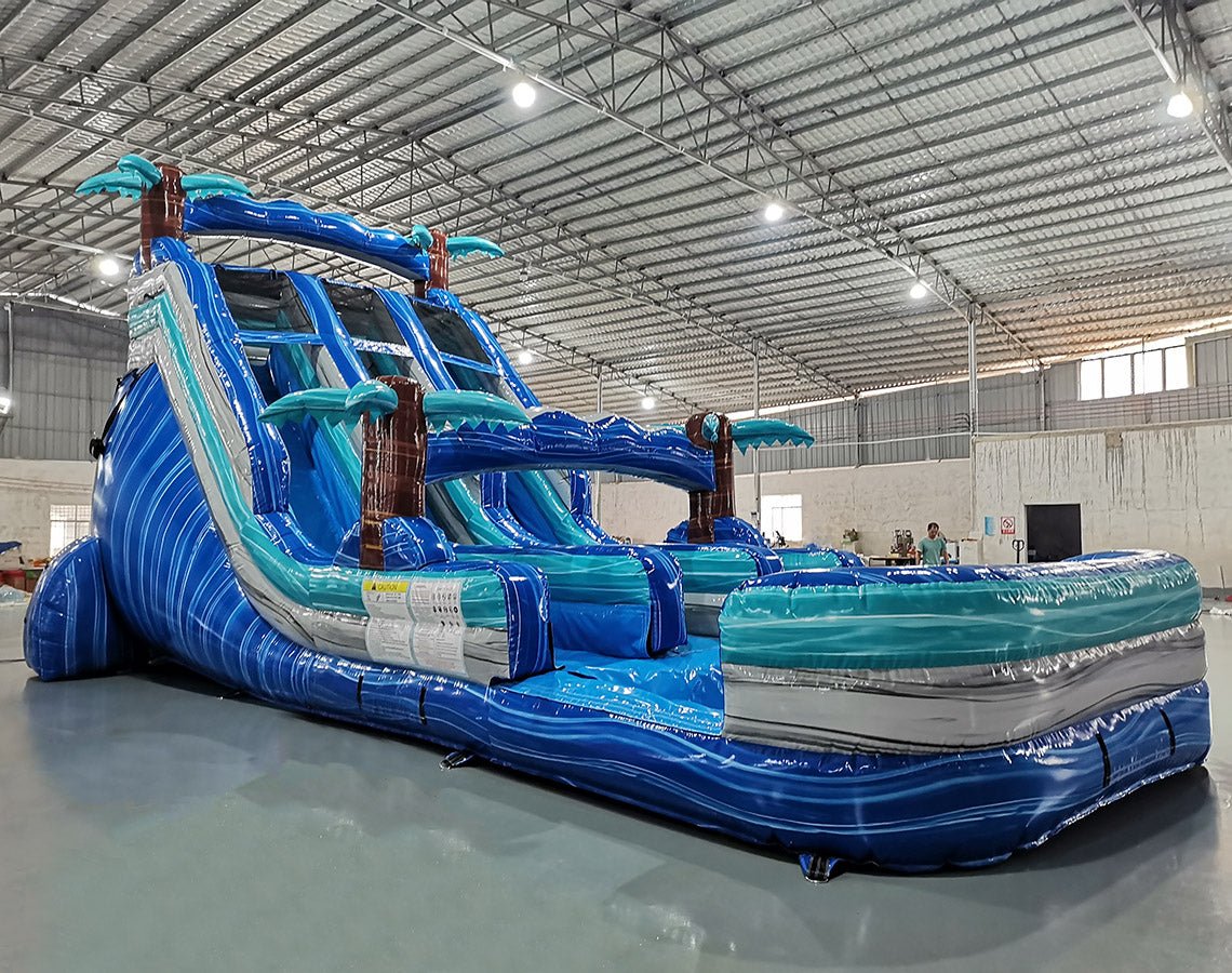 Angled view of 18ft Cayman Crush Center Climb water slide showcasing its full length, three blue sliding lanes, inflatable palm tree decorations, central climbing section, and extended turquoise splash pool in a large indoor warehouse.