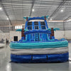 Front view of 18ft Cayman Crush Center Climb water slide showing three blue sliding lanes, inflatable palm trees, central climbing area with safety netting, and spacious turquoise splash pool in an indoor warehouse setting.