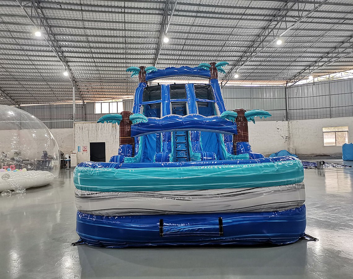 Front view of 18ft Cayman Crush Center Climb water slide showing three blue sliding lanes, inflatable palm trees, central climbing area with safety netting, and spacious turquoise splash pool in an indoor warehouse setting.