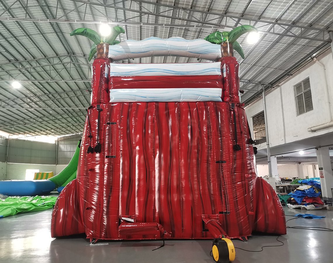 Rear view of 18ft Crimson Bay inflatable water slide showing red support structure, blue wave-patterned top, and green palm tree decorations, with a yellow air pump visible at the base.