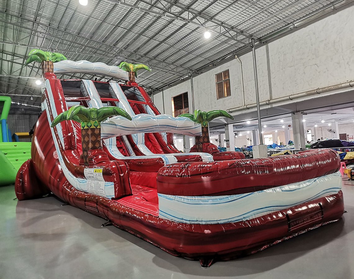 Front angled view of 18ft Crimson Bay water slide showcasing three red sliding lanes with blue wave patterns, green palm tree decorations, and central climbing structure in a spacious indoor facility.