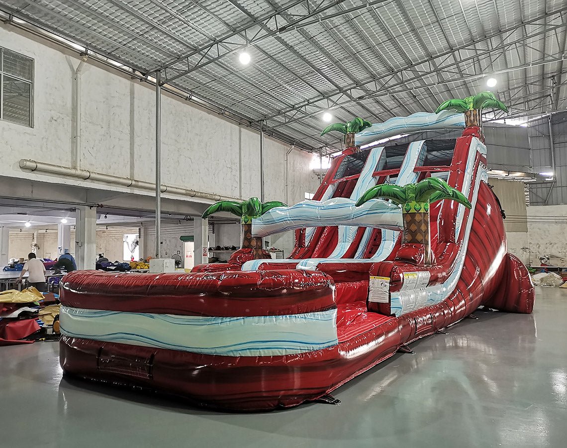 Large inflatable water slide with red base, blue wave patterns, and green palm tree decorations, featuring three sliding lanes and a central climbing structure, set up in an indoor warehouse.