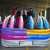 Front view of the 18ft Dino Duel inflatable water slide, highlighting its dual lanes in pink and blue, central climbing structure, dinosaur decorations, and inflatable palm trees, displayed in a large indoor space with other inflatables visible.