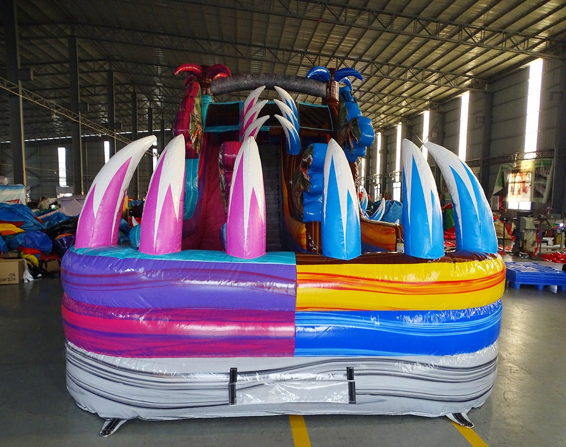 Front view of the 18ft Dino Duel inflatable water slide, highlighting its dual lanes in pink and blue, central climbing structure, dinosaur decorations, and inflatable palm trees, displayed in a large indoor space with other inflatables visible.