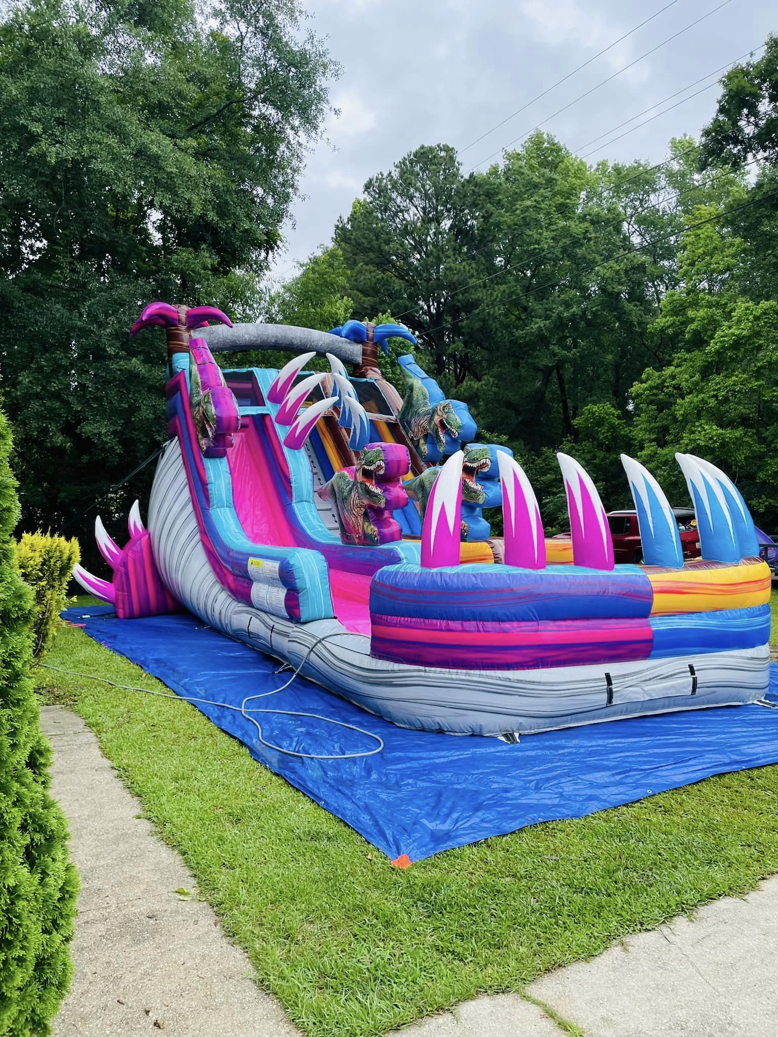 Side view of the 18ft Dino Duel water slide in an indoor warehouse, showcasing its colorful design with pink and blue lanes, dinosaur decorations, palm tree inflatables, and a grey base resembling rock formations.