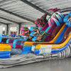 Large inflatable water slide featuring dinosaur themes, with two sliding lanes in pink and blue, inflatable palm trees, and multiple dinosaur figures positioned throughout the structure, set up in an indoor warehouse.