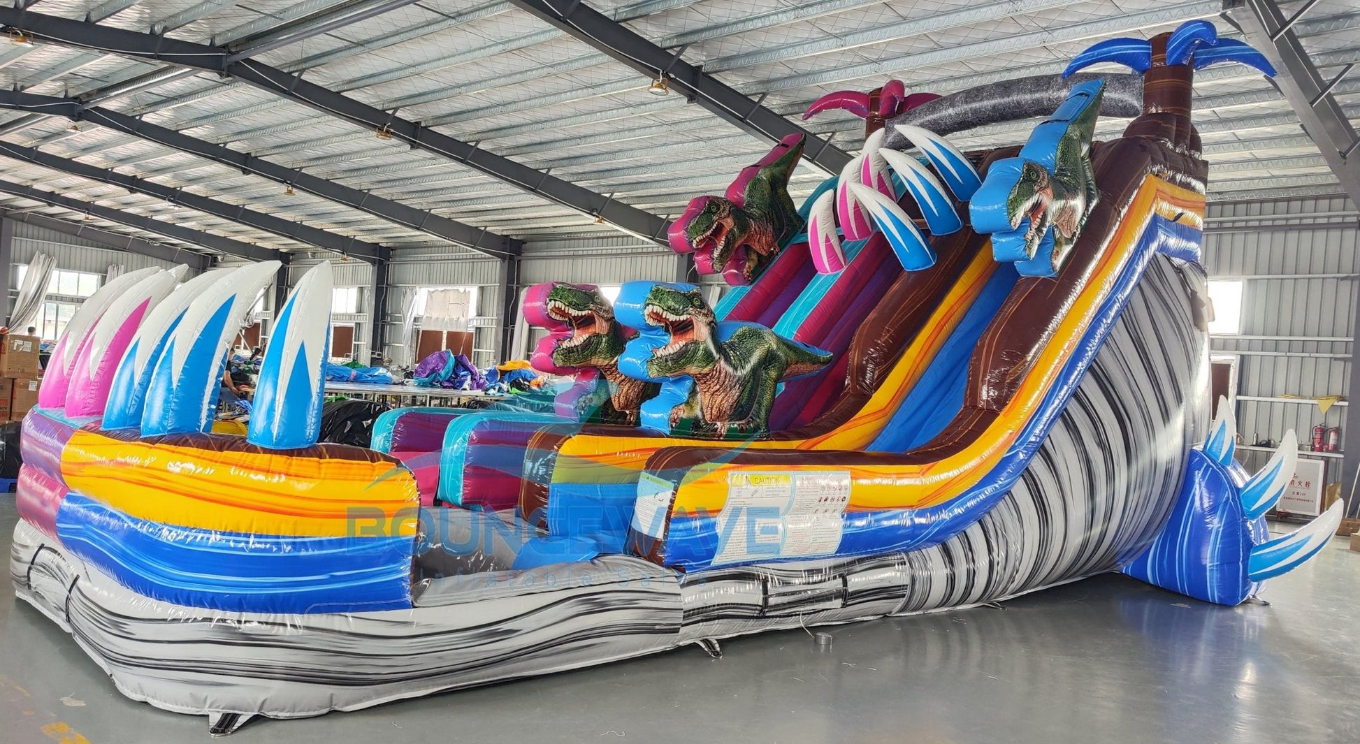 Large inflatable water slide featuring dinosaur themes, with two sliding lanes in pink and blue, inflatable palm trees, and multiple dinosaur figures positioned throughout the structure, set up in an indoor warehouse.