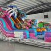 Full-length view of the 18ft Dino Duel Center Climb water slide, showcasing its colorful design with pink and blue lanes, dinosaur figures, palm trees, and a large splash pool area at the base, set up in an indoor facility.