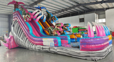 Full-length view of the 18ft Dino Duel Center Climb water slide, showcasing its colorful design with pink and blue lanes, dinosaur figures, palm trees, and a large splash pool area at the base, set up in an indoor facility.