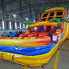 Full-length view of the 18ft Fiesta Fire inflatable water slide, showcasing its three sliding lanes, central climbing structure, palm tree decorations, and large splash pool area, set up in a spacious indoor facility with other inflatables visible.