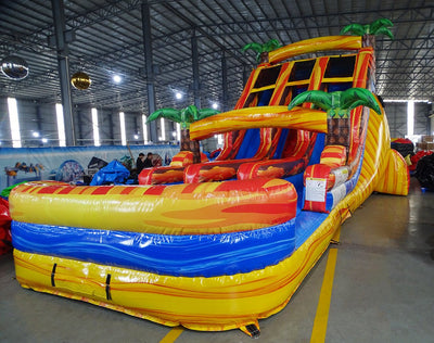 Full-length view of the 18ft Fiesta Fire inflatable water slide, showcasing its three sliding lanes, central climbing structure, palm tree decorations, and large splash pool area, set up in a spacious indoor facility with other inflatables visible.