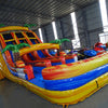 Side angle view of 18ft Fiesta Fire water slide showing vibrant red, yellow, and blue colors, inflatable palm trees, and a large splash pool area at the base, surrounded by other inflatables in a warehouse setting.