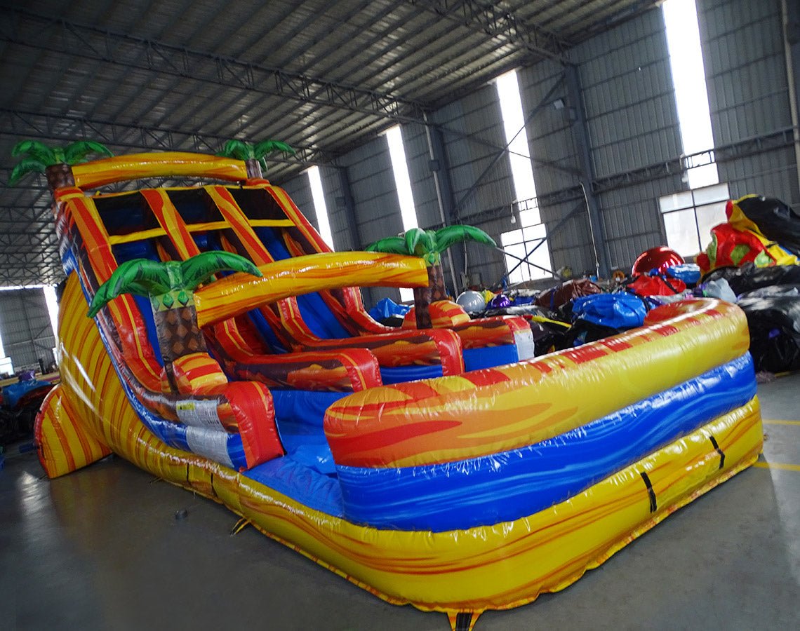 Side angle view of 18ft Fiesta Fire water slide showing vibrant red, yellow, and blue colors, inflatable palm trees, and a large splash pool area at the base, surrounded by other inflatables in a warehouse setting.