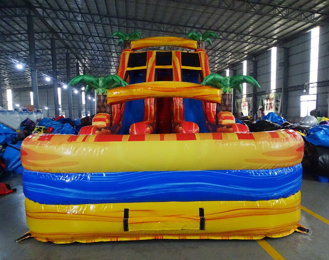 Front view of large inflatable water slide with tropical theme, featuring three red and yellow sliding lanes, palm tree decorations, and a central climbing structure, set up in an indoor warehouse.