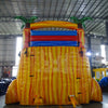 Large inflatable water slide with tropical theme, featuring three red, yellow, and blue sliding lanes, palm tree decorations, and a central climbing structure, set up in an indoor warehouse surrounded by other inflatables and equipment.