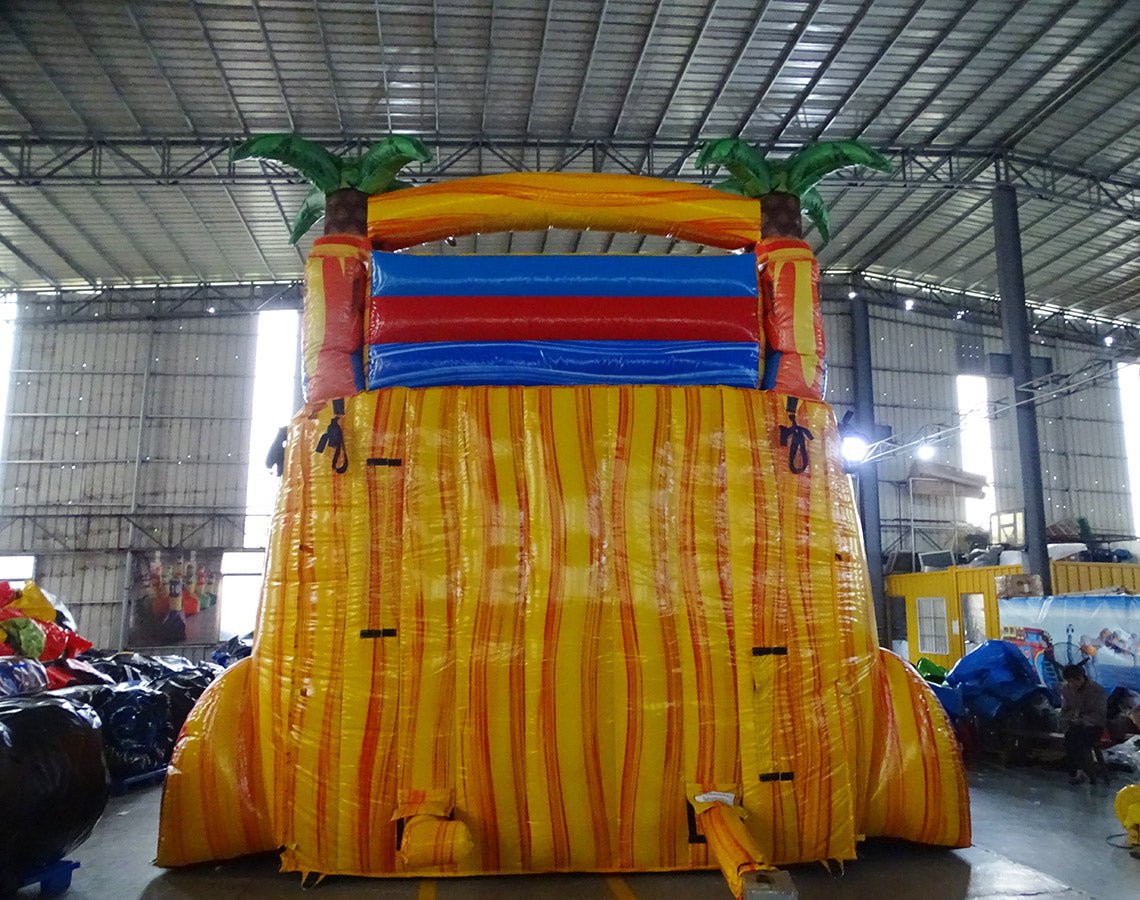 Large inflatable water slide with tropical theme, featuring three red, yellow, and blue sliding lanes, palm tree decorations, and a central climbing structure, set up in an indoor warehouse surrounded by other inflatables and equipment.