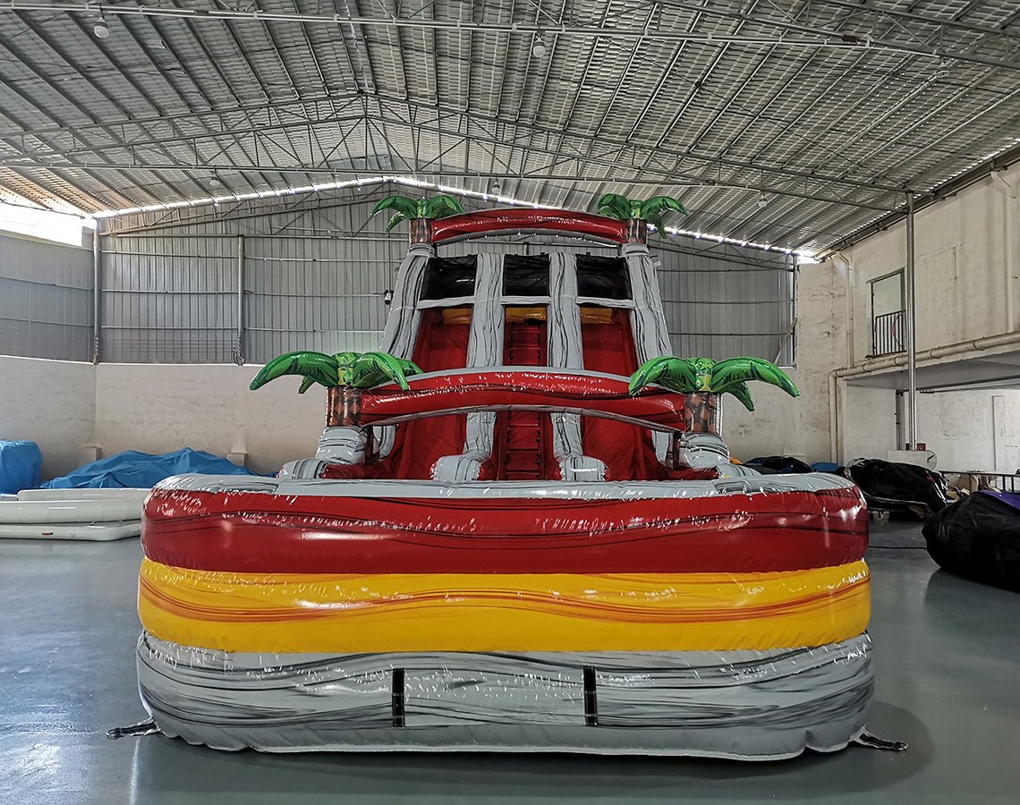 Front view of 18ft Fire Island inflatable water slide featuring red, yellow, and gray color scheme, three sliding lanes, inflatable palm trees, and a central climbing area, displayed in a large indoor warehouse.