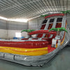 Large inflatable water slide with tropical theme, featuring three red, yellow, and gray sliding lanes, palm tree decorations, and a central climbing structure, set up in an indoor warehouse with metal roofing.