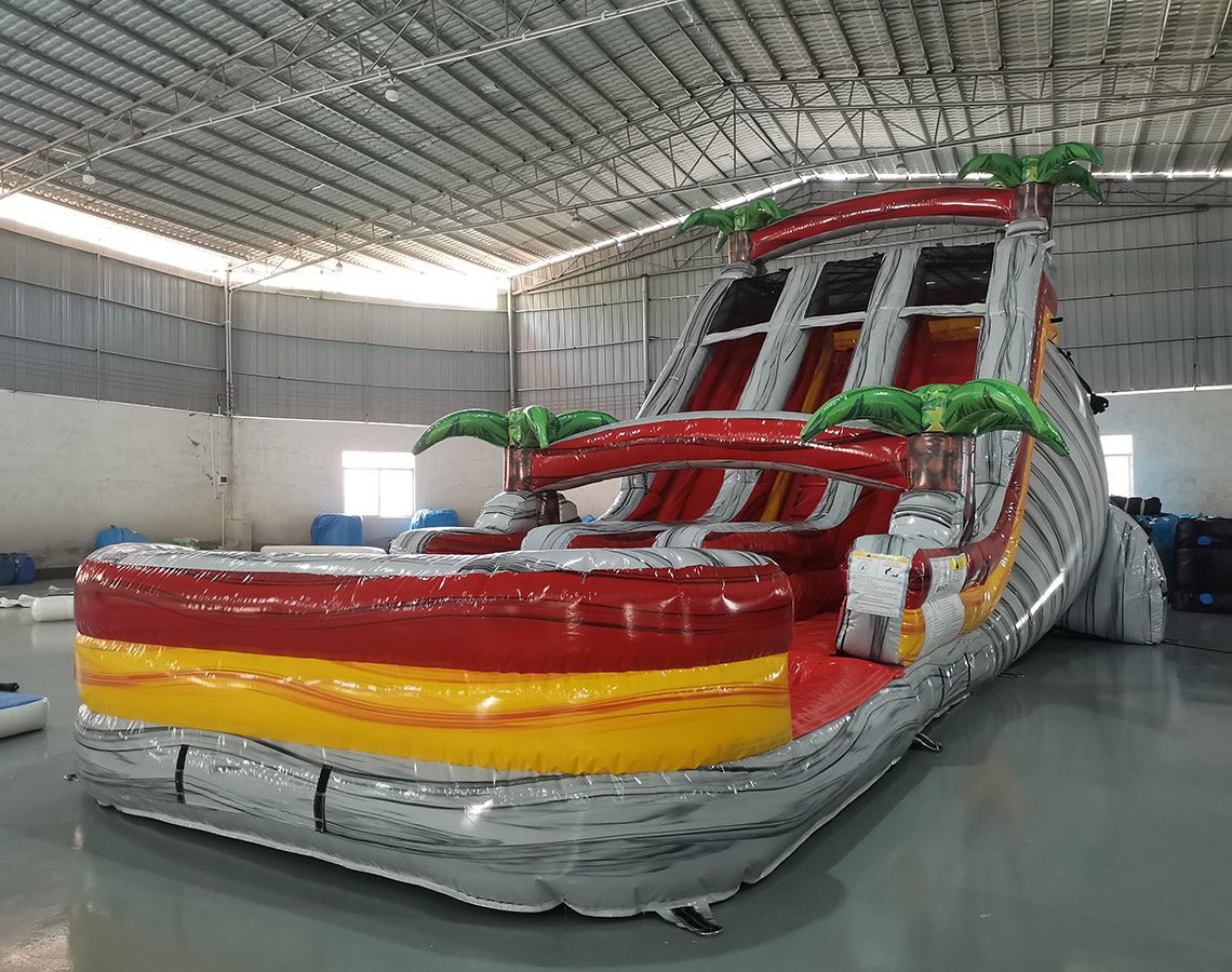 Large inflatable water slide with tropical theme, featuring three red, yellow, and gray sliding lanes, palm tree decorations, and a central climbing structure, set up in an indoor warehouse with metal roofing.