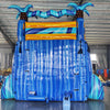 Large inflatable water slide with tropical theme, featuring three blue sliding lanes, palm tree decorations, and a central climbing structure, set up in an indoor warehouse with metal roofing and other equipment visible.