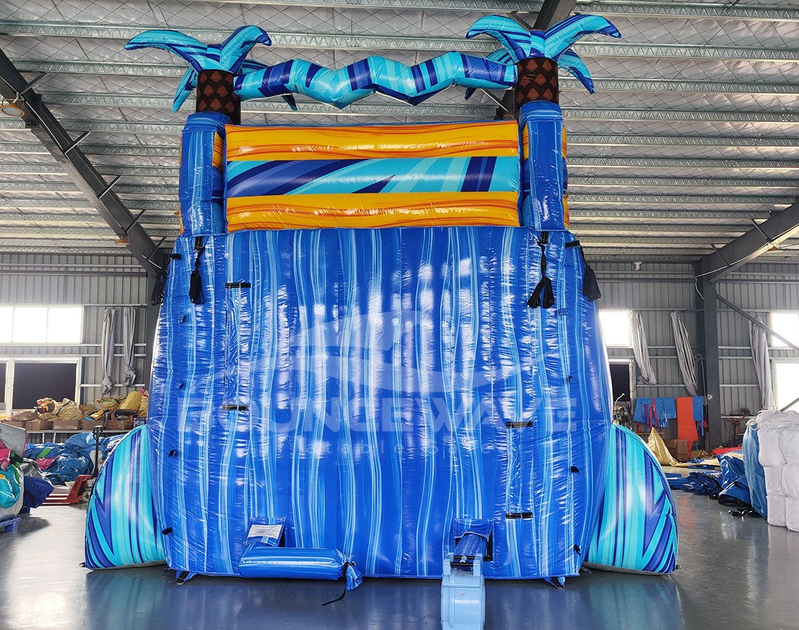 Large inflatable water slide with tropical theme, featuring three blue sliding lanes, palm tree decorations, and a central climbing structure, set up in an indoor warehouse with metal roofing and other equipment visible.