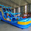 Large inflatable water slide with tropical theme, featuring three blue sliding lanes, palm tree decorations, and a central climbing structure, set up in an indoor warehouse with metal roofing and other equipment visible.