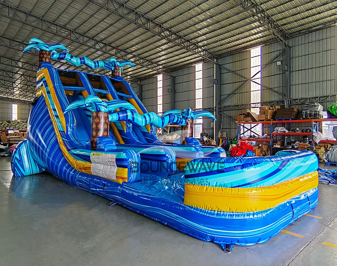 Large inflatable water slide with tropical theme, featuring three blue sliding lanes, palm tree decorations, and a central climbing structure, set up in an indoor warehouse with metal roofing and other equipment visible.