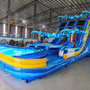 Angled view of the 18ft Flashpoint Falls Center Climb water slide, showcasing its three sliding lanes, central climbing structure, palm tree decorations, and large splash pool area, set up in a spacious indoor facility with holiday decorations visible.