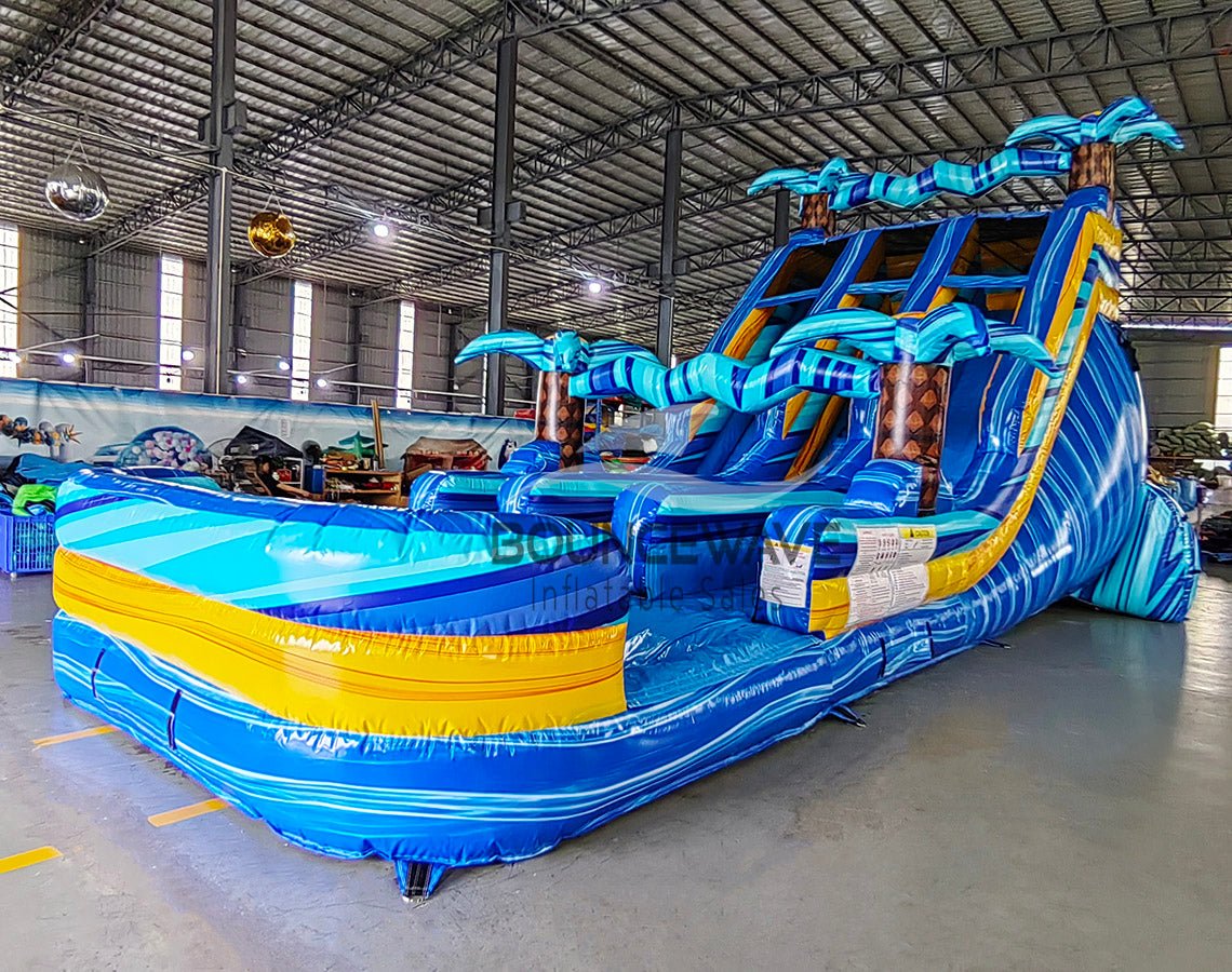 Angled view of the 18ft Flashpoint Falls Center Climb water slide, showcasing its three sliding lanes, central climbing structure, palm tree decorations, and large splash pool area, set up in a spacious indoor facility with holiday decorations visible.