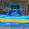 Front view of 18ft Flashpoint Falls inflatable water slide featuring blue and yellow color scheme, three sliding lanes, inflatable palm trees, and a central climbing area, displayed in a large indoor warehouse with other inflatables in the background.