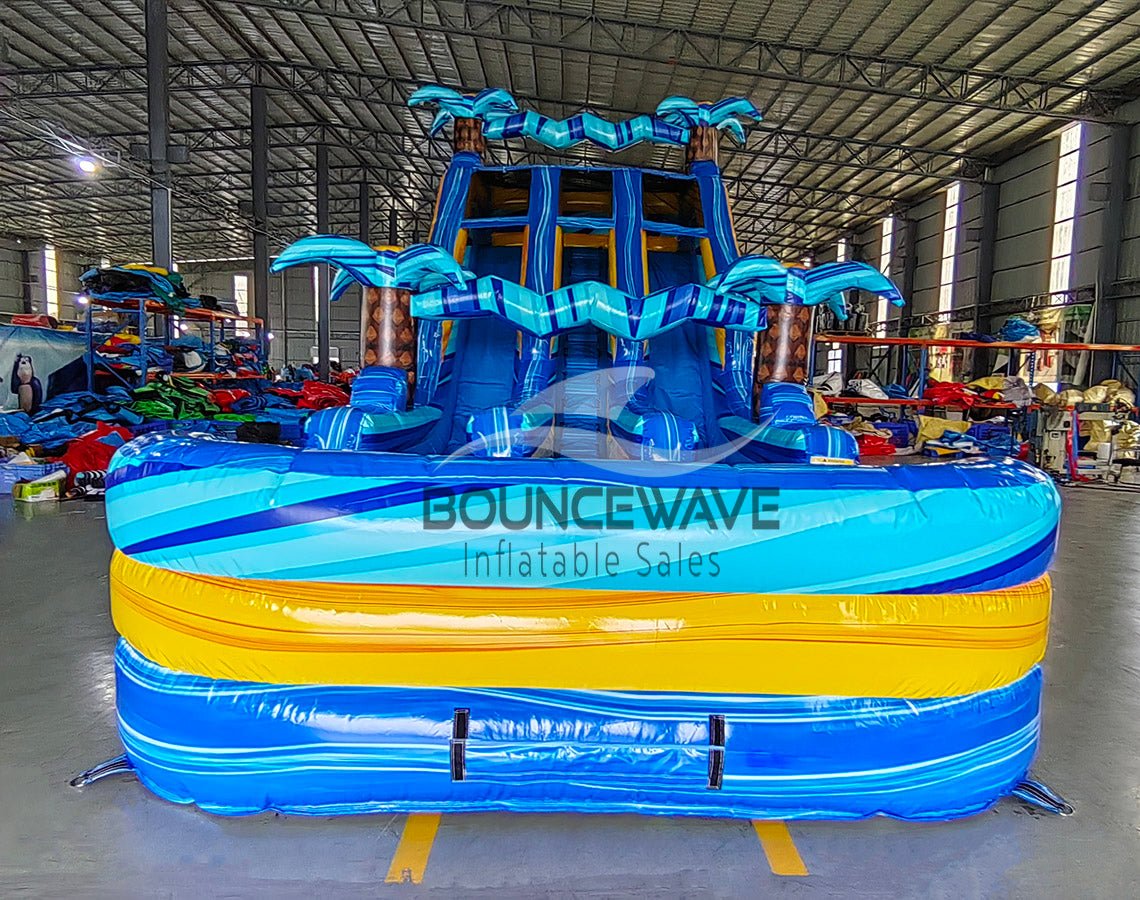 Front view of 18ft Flashpoint Falls inflatable water slide featuring blue and yellow color scheme, three sliding lanes, inflatable palm trees, and a central climbing area, displayed in a large indoor warehouse with other inflatables in the background.