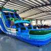 Angled view of the 18ft Island Drop inflatable water slide, showcasing its blue sliding lanes, palm tree decorations, central climbing structure, and large splash pool area, set up in a warehouse with metal roofing visible.