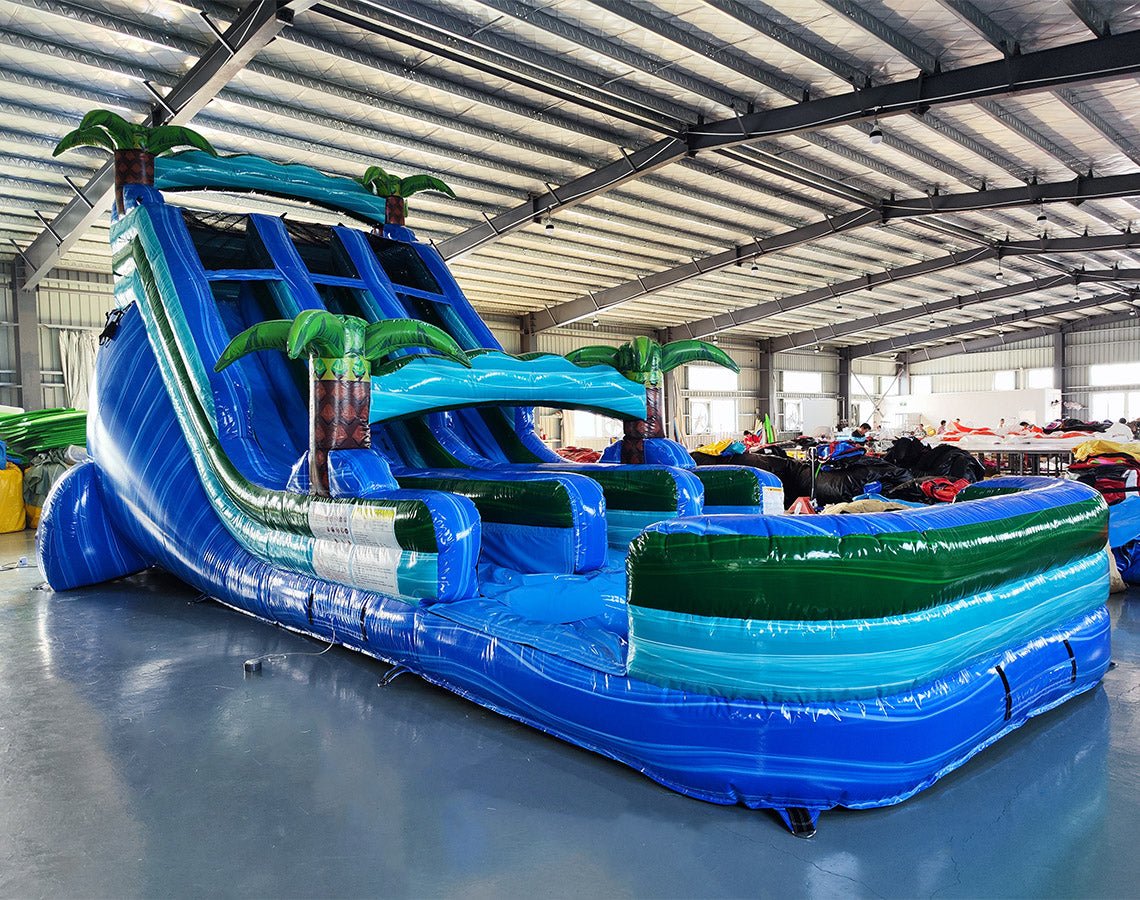 Angled view of the 18ft Island Drop inflatable water slide, showcasing its blue sliding lanes, palm tree decorations, central climbing structure, and large splash pool area, set up in a warehouse with metal roofing visible.