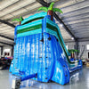 Large inflatable water slide with tropical theme, featuring three blue sliding lanes, palm tree decorations, and a central climbing structure, set up in an indoor warehouse with other inflatables and equipment visible in the background.