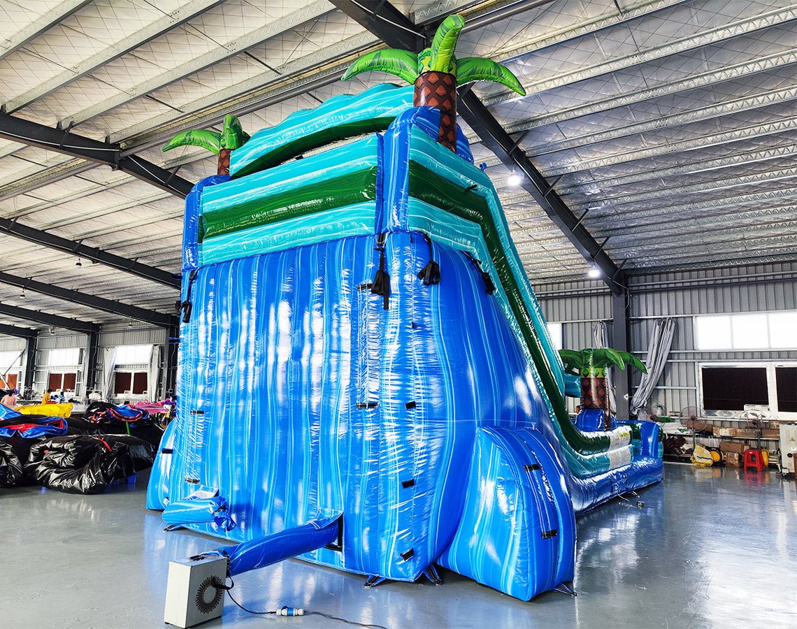 Large inflatable water slide with tropical theme, featuring three blue sliding lanes, palm tree decorations, and a central climbing structure, set up in an indoor warehouse with other inflatables and equipment visible in the background.