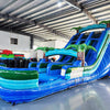 Side view of 18ft Island Drop water slide in an indoor warehouse, showing vibrant blue lanes, inflatable palm trees, and a large splash pool area with green and blue color scheme.