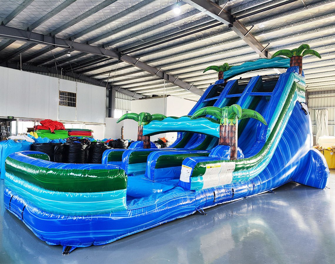 Side view of 18ft Island Drop water slide in an indoor warehouse, showing vibrant blue lanes, inflatable palm trees, and a large splash pool area with green and blue color scheme.