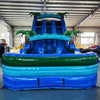 Front view of 18ft Island Drop Center Climb water slide, displaying three blue sliding lanes, inflatable palm trees, and a spacious splash pool with green borders, set up in an indoor facility with other inflatables visible.