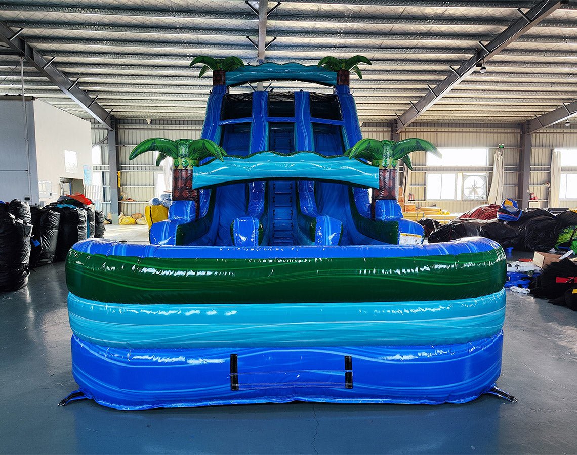 Front view of 18ft Island Drop Center Climb water slide, displaying three blue sliding lanes, inflatable palm trees, and a spacious splash pool with green borders, set up in an indoor facility with other inflatables visible.